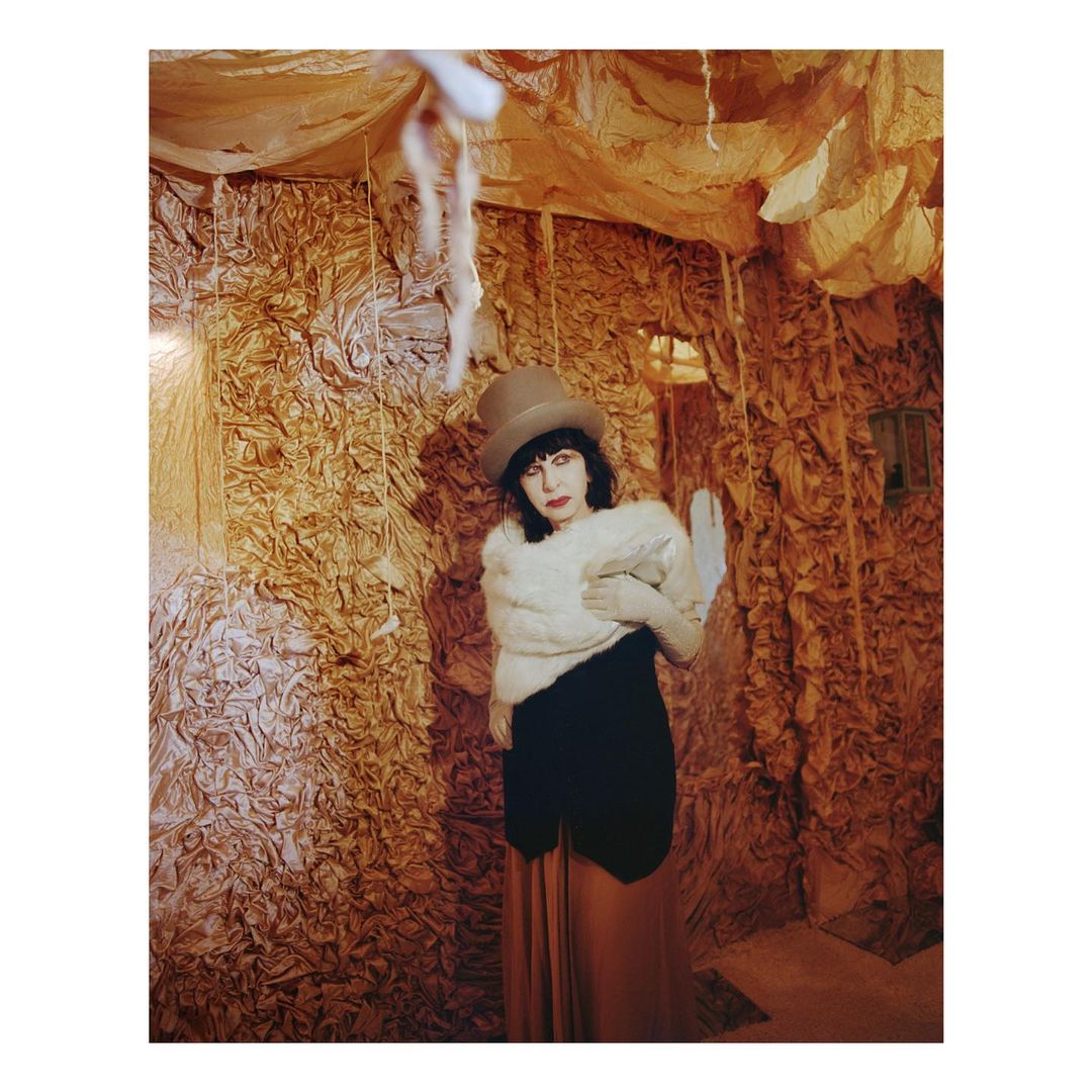 Artistic portrait of an adult female in a hat and shawl posing in an elaborate, textured, warm-toned indoor setting.