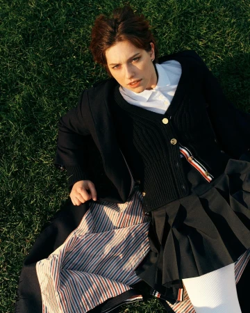 A woman with short brown hair wearing a dark pleated skirt and cardigan lying on vibrant green grass.