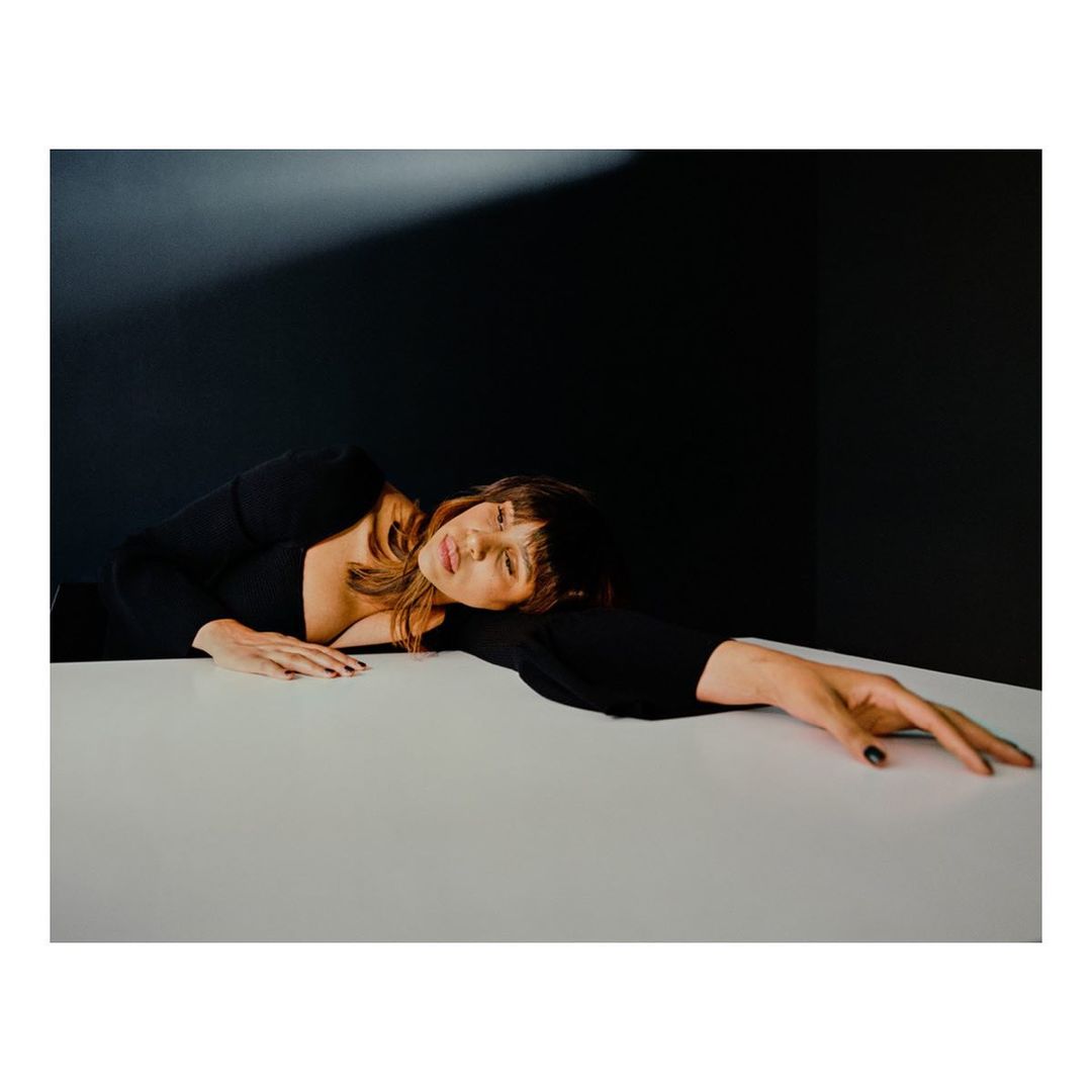 Adult woman with bangs resting on a white table wearing a black long-sleeved shirt, against a dark background with light source.