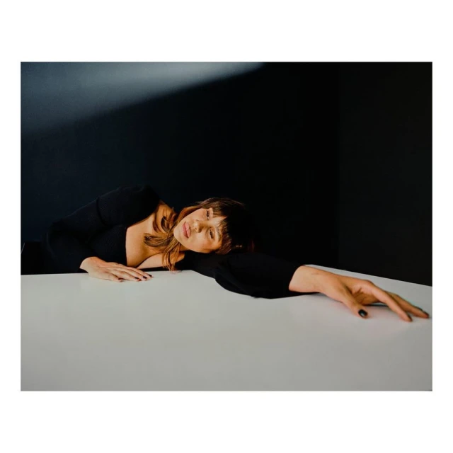 Adult woman with bangs resting on a white table wearing a black long-sleeved shirt, against a dark background with light source.