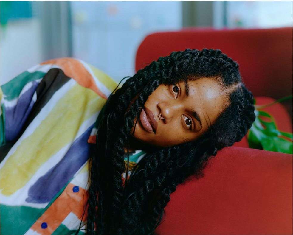 Portrait of a Black woman with braided hair, lying on a red sofa, wearing a colorful striped shirt. Natural lighting illuminates the scene.