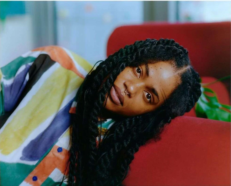 Portrait of a Black woman with braided hair, lying on a red sofa, wearing a colorful striped shirt. Natural lighting illuminates the scene.