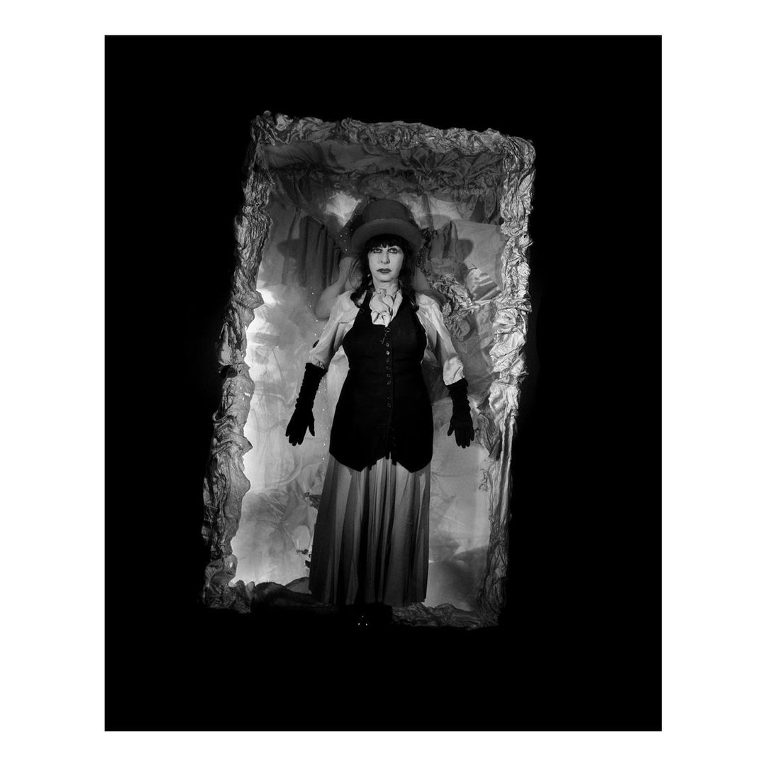 Monochrome portrait of a woman in a top hat and vest standing in a decorative frame with an ornate backdrop, set against a black background.