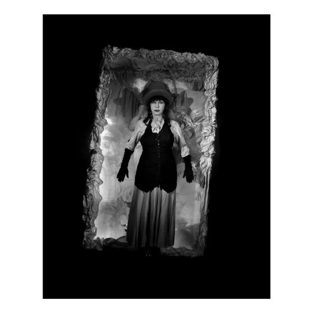 Monochrome portrait of a woman in a top hat and vest standing in a decorative frame with an ornate backdrop, set against a black background.