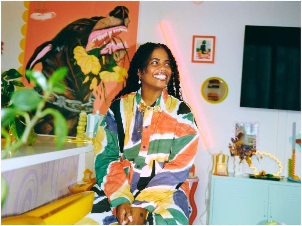 Smiling woman in colorful shirt poses in an eclectic interior setting with paintings, plants, and a light-blue storage cabinet.