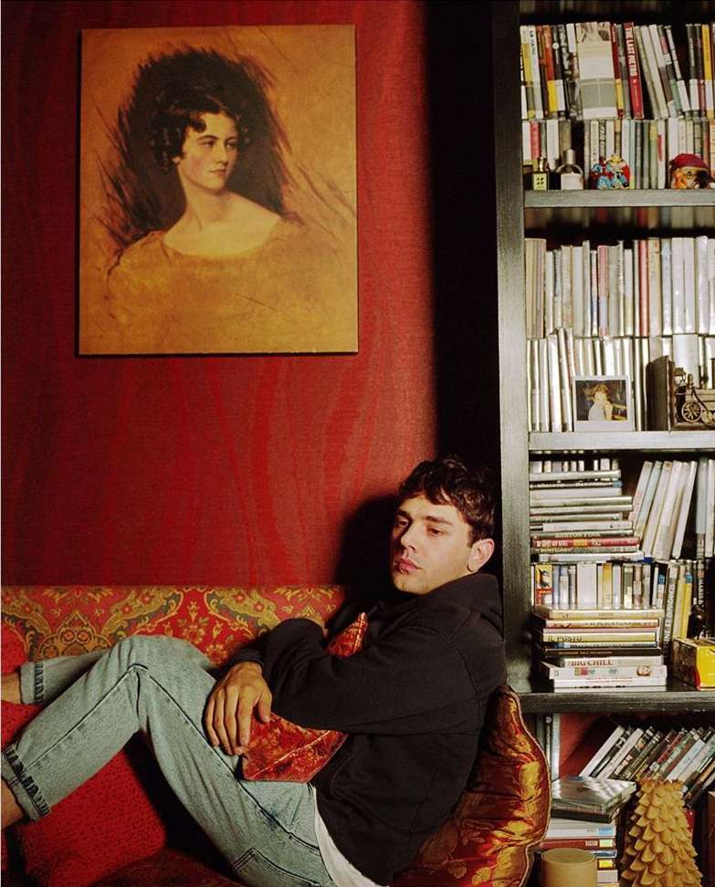 Adult male lounges on an ornate couch in a red-walled room with a bookcase and portrait in the background. Relaxed indoor setting.