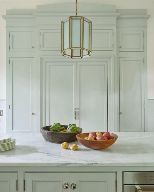 A bright kitchen interior showcases a marble countertop with wooden bowls filled with potatoes and artichokes, beneath a modern pendant light.