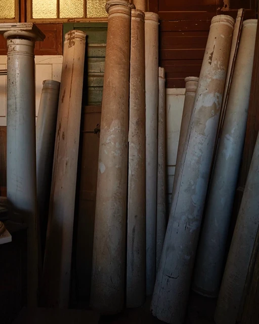 Group of large, weathered architectural columns stacked indoors with a window in the background providing soft, diffused lighting.