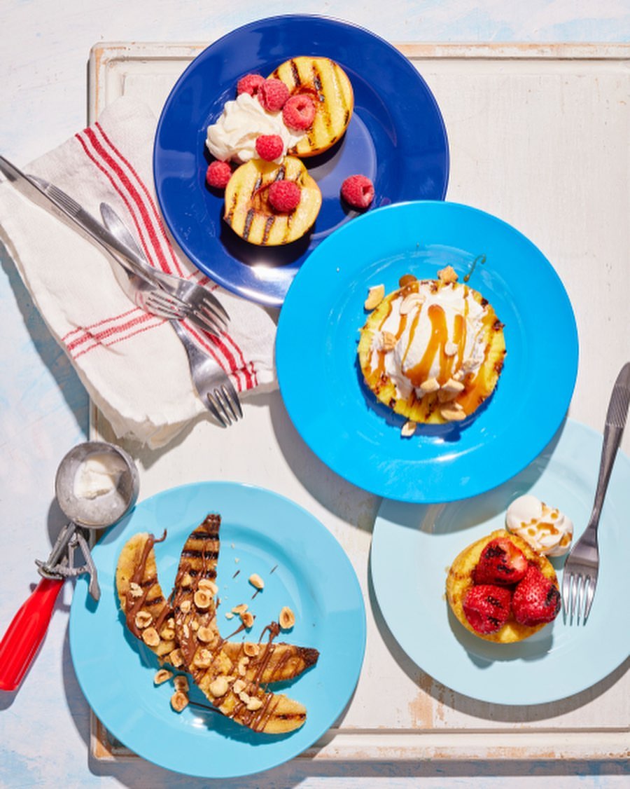 Overhead shot of four colorful plates, each holding a grilled fruit dessert with toppings like ice cream, raspberries, and caramel.