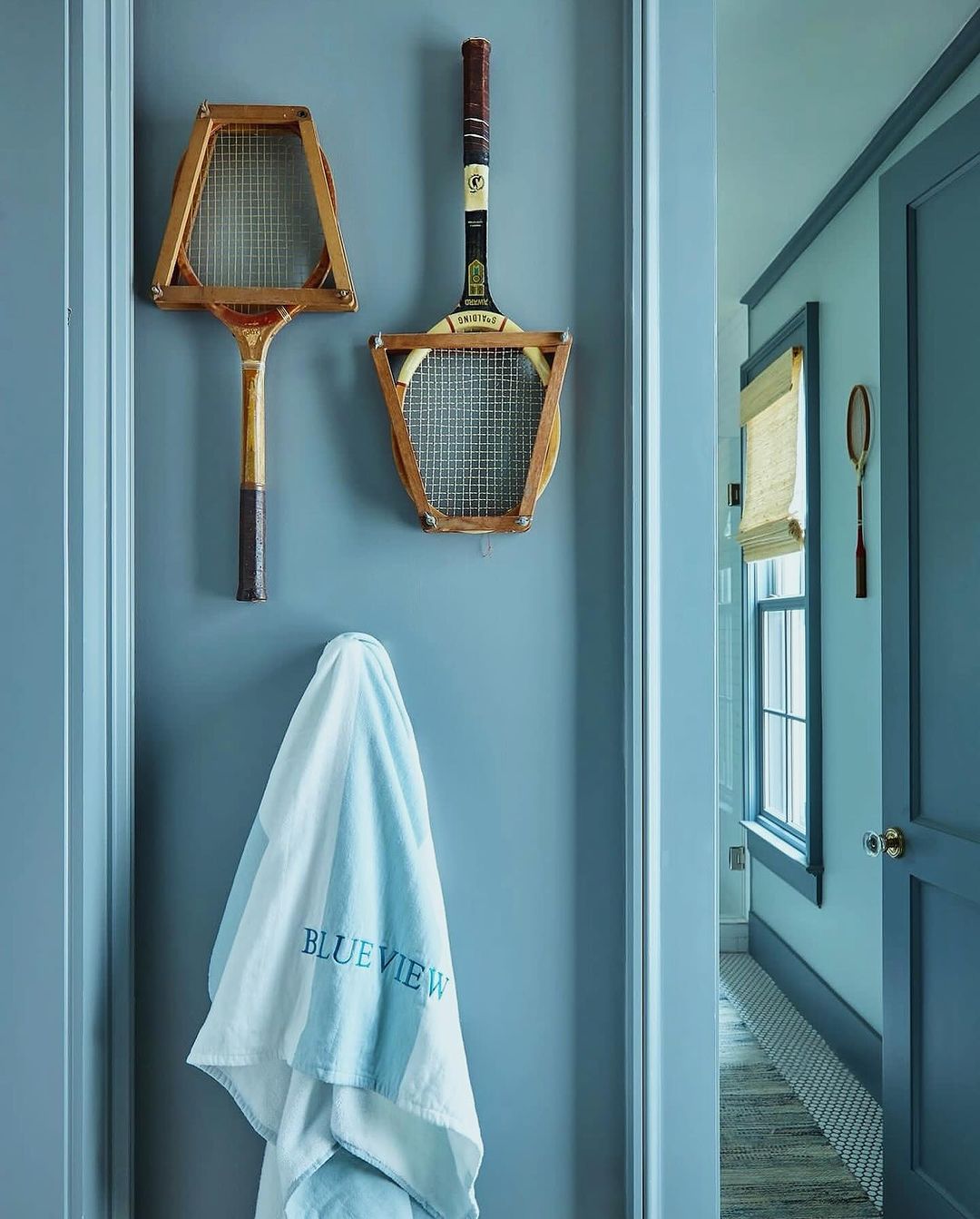 Two vintage wooden tennis rackets hang on a blue wall with a blue and white towel hanging below them in an indoor setting.