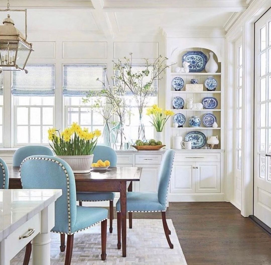 Dining room interior with a wooden table, blue upholstered chairs, yellow daffodils, and built-in shelving with blue and white china.