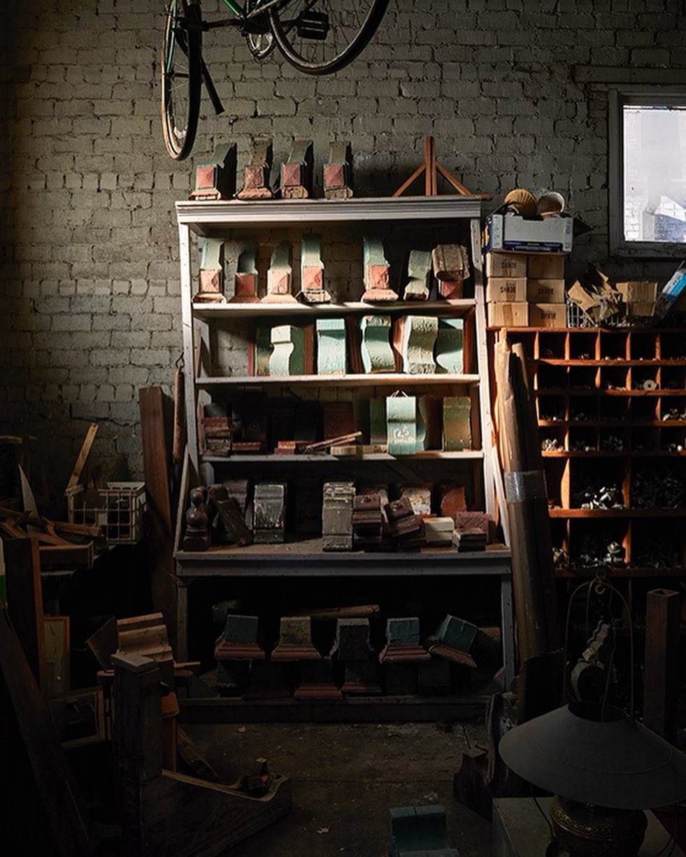 Vintage shelving unit displays assorted wooden objects inside a workshop under a hanging bicycle frame against a brick wall.