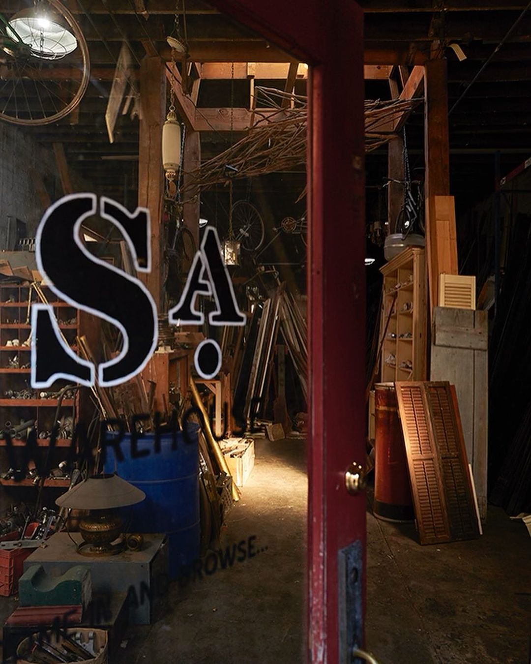 The interior of a cluttered retail space is viewed through a window with signage. Lighting illuminates the room, highlighting the variety of objects.