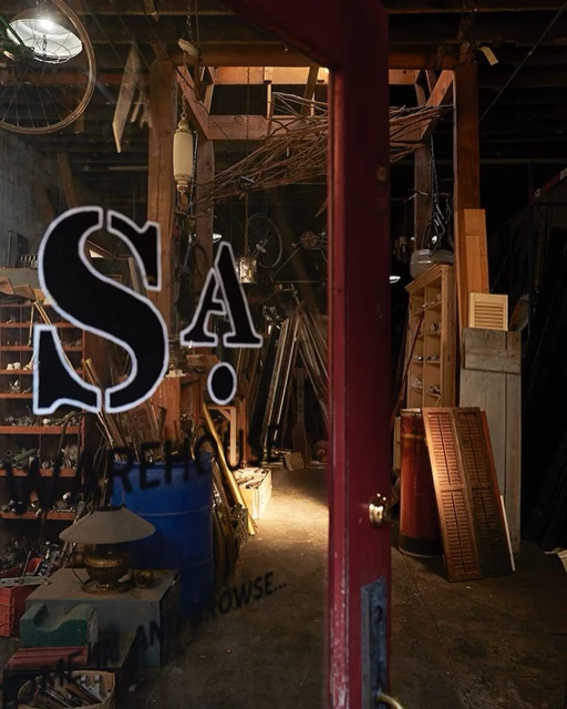 The interior of a cluttered retail space is viewed through a window with signage. Lighting illuminates the room, highlighting the variety of objects.