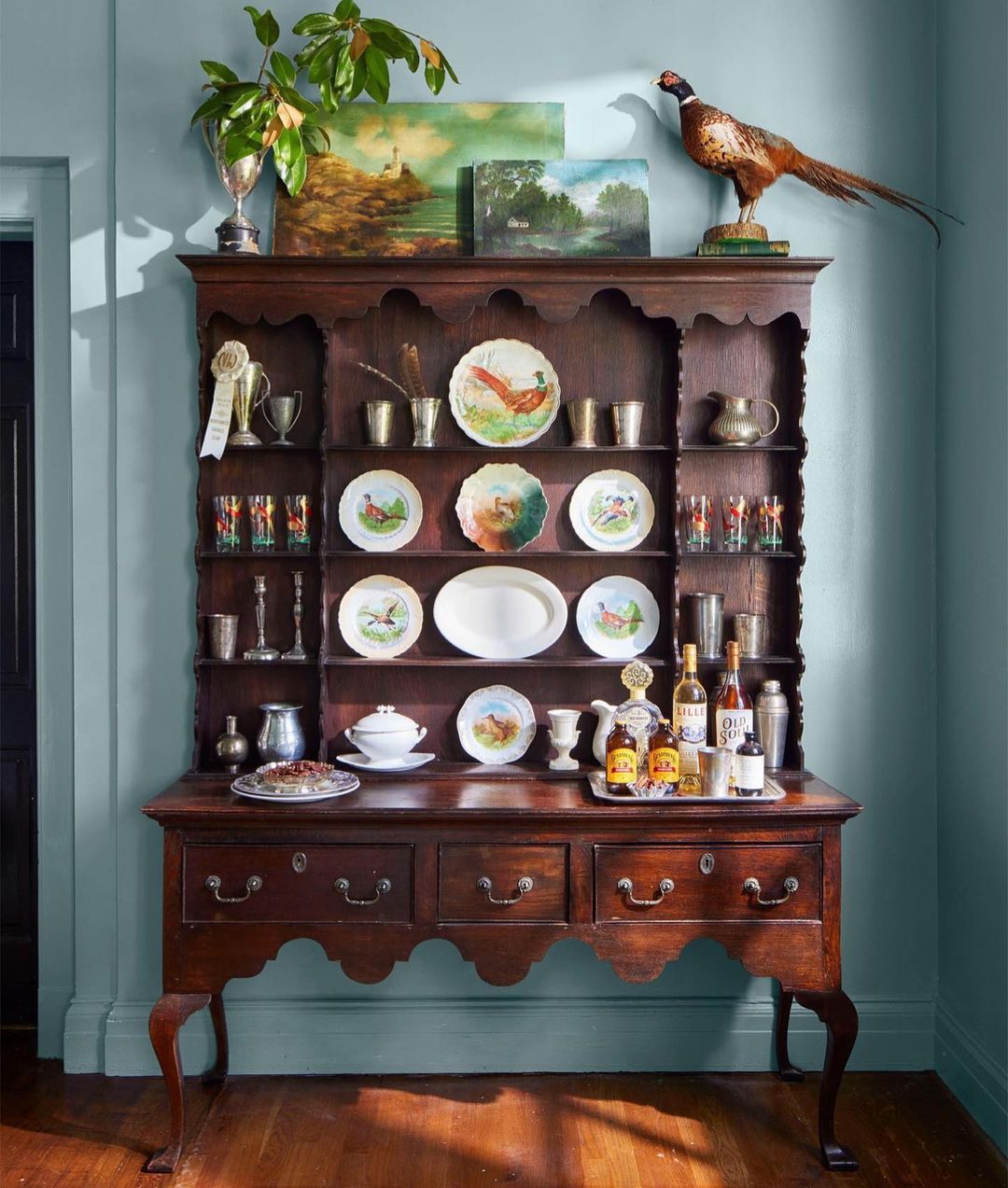Antique hutch filled with pheasant themed dishware and paintings, silver trophies, and glassware against a blue painted wall.