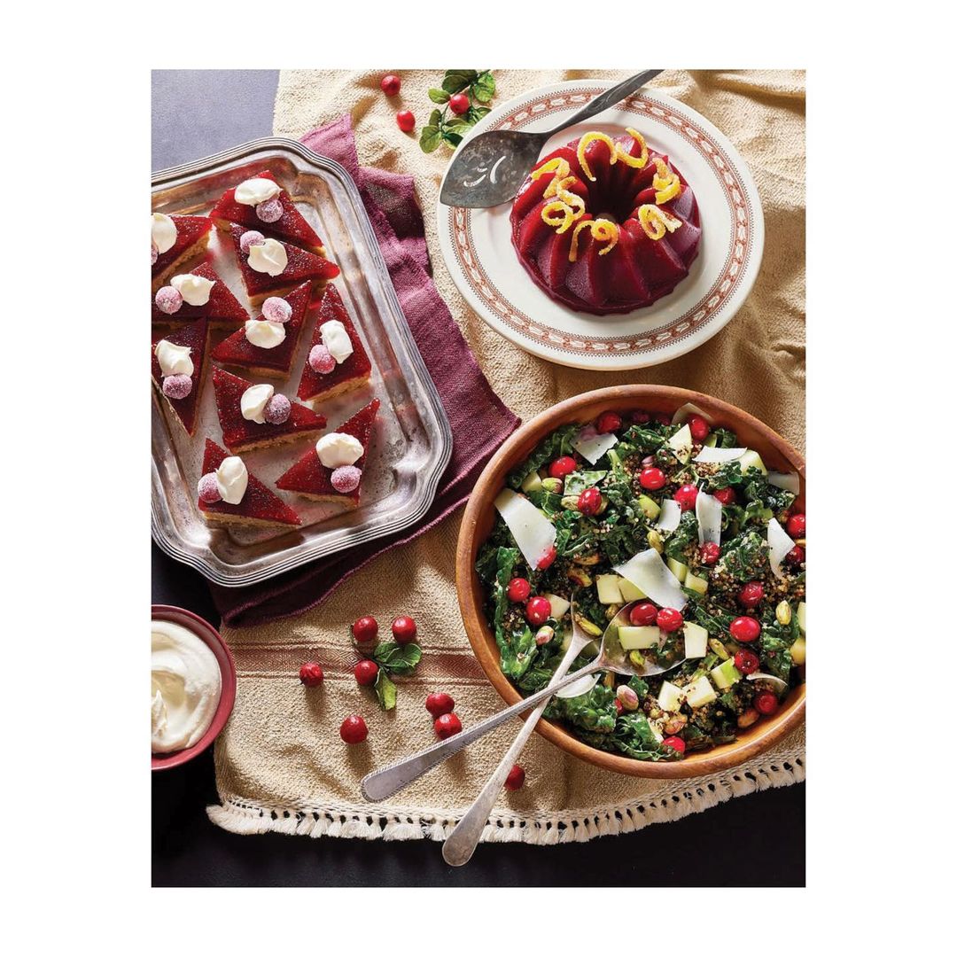 Overhead shot of a table with various dishes, including cranberry salad, cake, and cheesecake bars, all arranged on a beige tablecloth.
