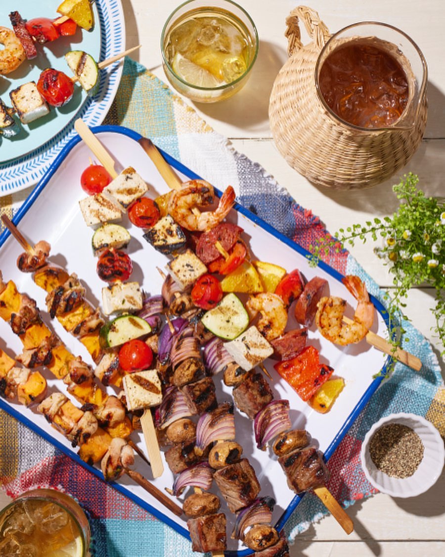 Grilled skewers of vegetables and meats, served on a white tray, with refreshing summer beverages and picnic cloth in background.