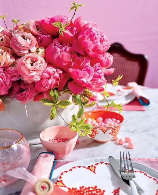 Close-up of a floral arrangement featuring pink peonies and roses in a white vase on a table with pink accents and tableware.