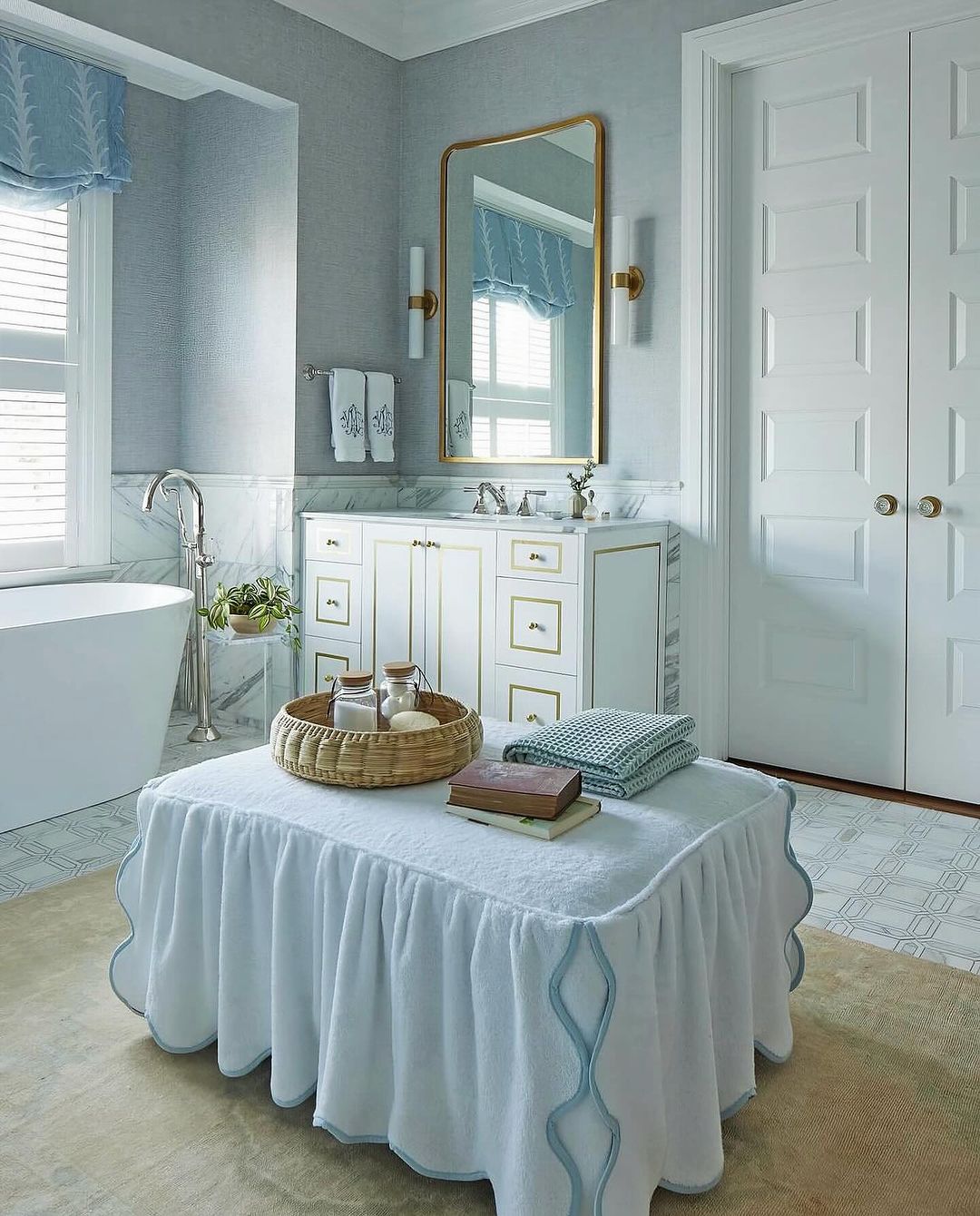 Elegant bathroom interior featuring a white vanity, gold mirror, and a skirted ottoman with towels and accessories in a bright, clean space.