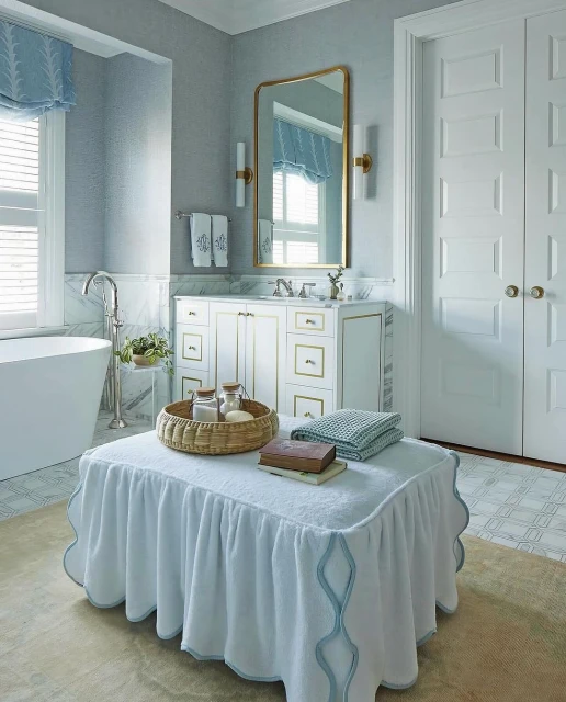 Elegant bathroom interior featuring a white vanity, gold mirror, and a skirted ottoman with towels and accessories in a bright, clean space.