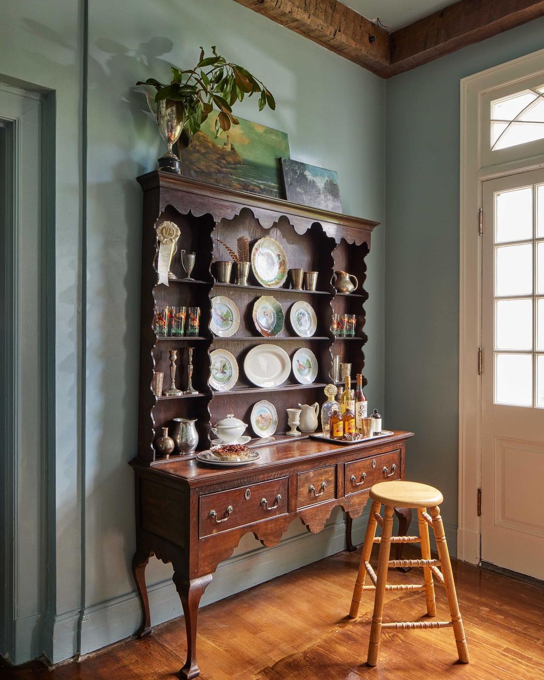 Dark wood hutch cabinet with displayed plates and barware in an interior with light blue walls and a wooden stool.