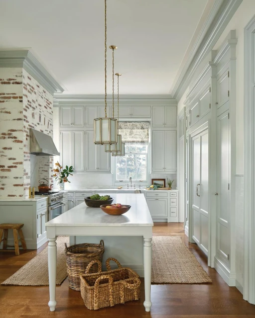 Bright kitchen with white cabinets, marble countertops, hardwood floors, brick accent wall, island with fruit bowls, and pendant lighting.
