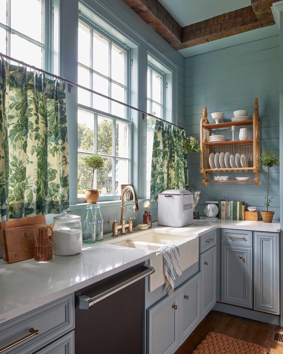 Well-lit kitchen interior showcasing light blue cabinetry, marble countertops, a farmhouse sink, and floral curtains covering windows.