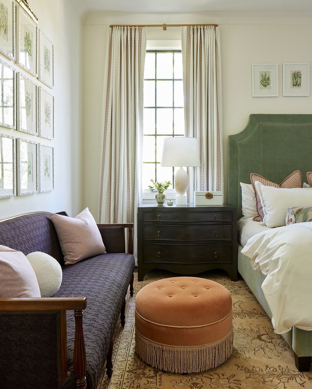 Bedroom interior with a green velvet headboard bed, dark wood chest, upholstered ottoman, settee, framed artwork, and patterned curtains.
