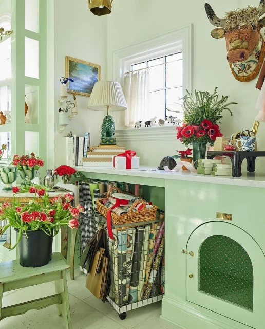 Interior home office with mint green desk and walls, featuring a floral bouquet, wrapped gifts, and organized wrapping paper supplies.