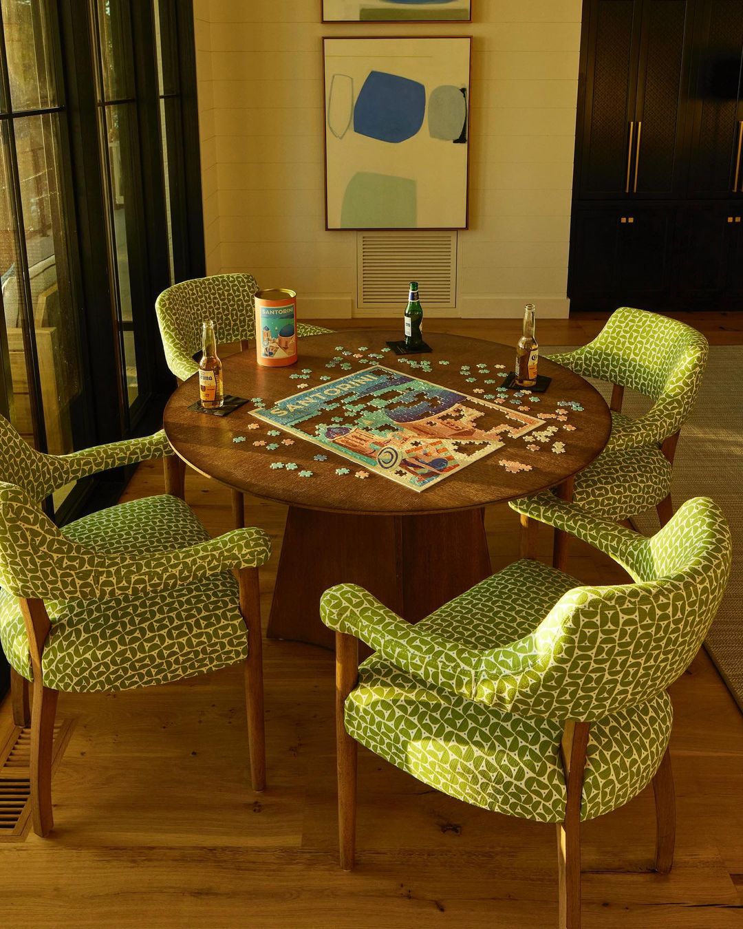 Round table with a jigsaw puzzle and surrounded by four armchairs featuring a green and white geometric pattern inside a living room.