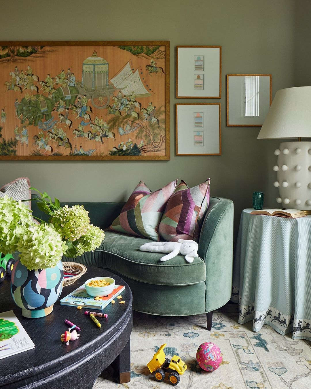 Living room interior with green curved sofa, floral decorations, artwork, coffee table with kids' items, and patterned rug.