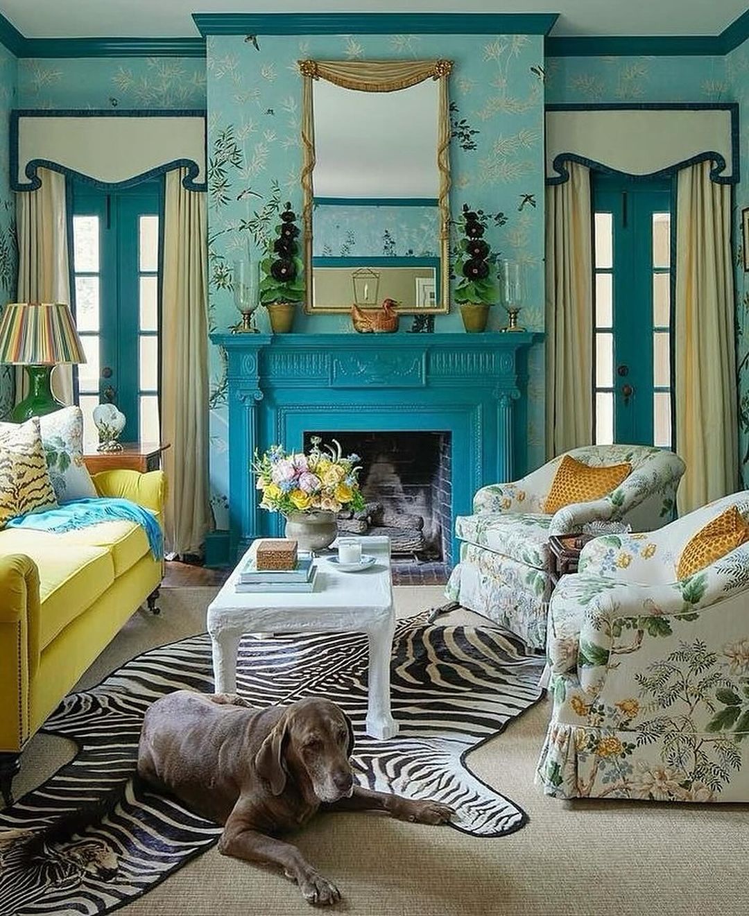 Living room interior featuring a dog relaxing on a zebra-print rug, a yellow sofa, armchairs, a fireplace, and blue patterned wallpaper.