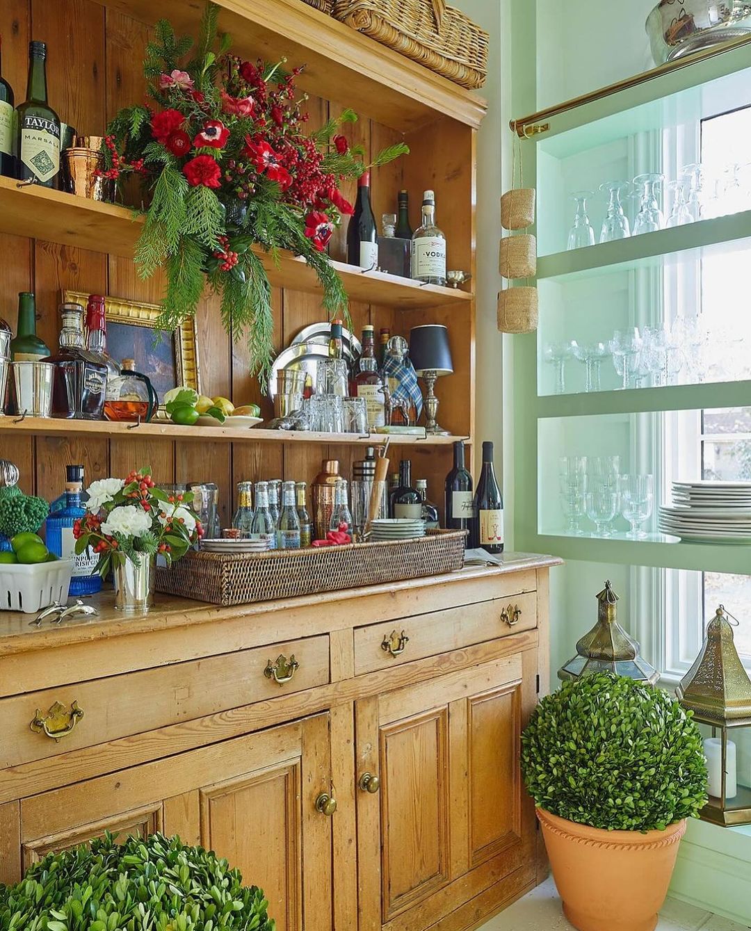 Home bar setup featuring a wooden hutch filled with glassware and bottles, decorated with floral arrangements and greenery, illuminated by natural light.