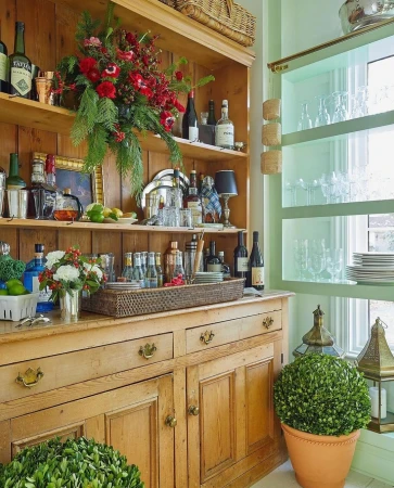 Home bar setup featuring a wooden hutch filled with glassware and bottles, decorated with floral arrangements and greenery, illuminated by natural light.