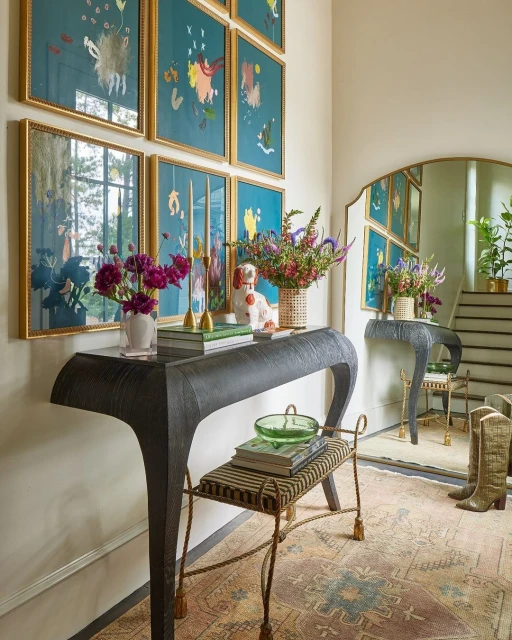Eclectic entryway with a grey console table adorned with flower arrangements and books, a mirror, and paintings against a beige wall.