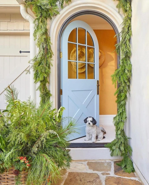 Small dog sits near a light blue arched door with evergreen garlands, and a stone walkway with a white paneled wall in the background.
