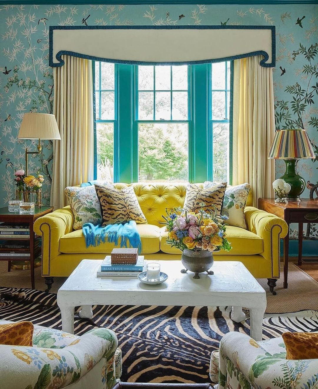 Bright living room interior with a yellow tufted sofa, floral armchairs, a white coffee table, and large window with garden view.