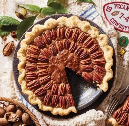 Overhead view of a baked pecan pie with a missing slice on a dark plate, surrounded by whole nuts and leaves.