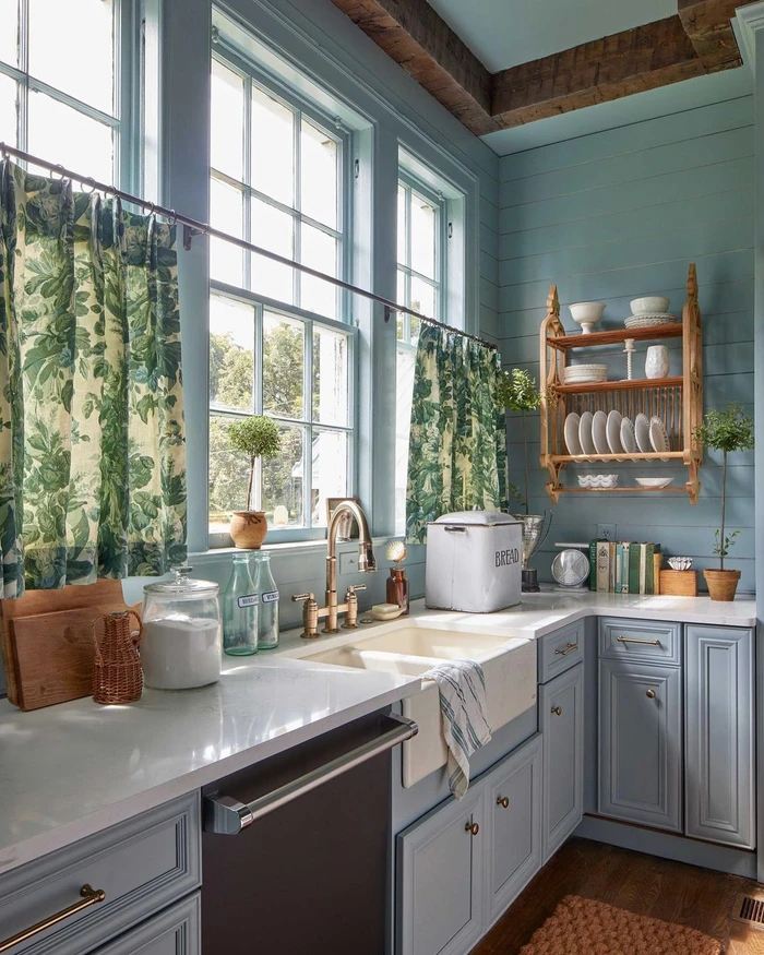 A brightly lit country kitchen featuring pale blue wooden paneled walls, white countertops, a large porcelain farmhouse sink, and green patterned cafe curtains hanging over tall multi-pane windows.