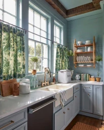 A brightly lit country kitchen featuring pale blue wooden paneled walls, white countertops, a large porcelain farmhouse sink, and green patterned cafe curtains hanging over tall multi-pane windows.