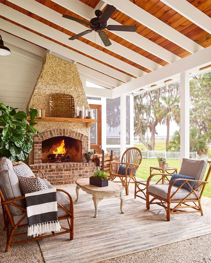 Covered outdoor patio with a large stone fireplace, wooden beam ceiling, rattan armchairs, sofa, and a lit fire.
