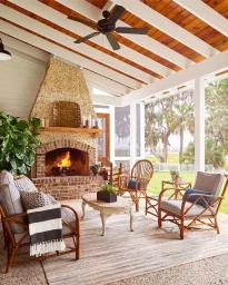 Covered outdoor patio with a large stone fireplace, wooden beam ceiling, rattan armchairs, sofa, and a lit fire.