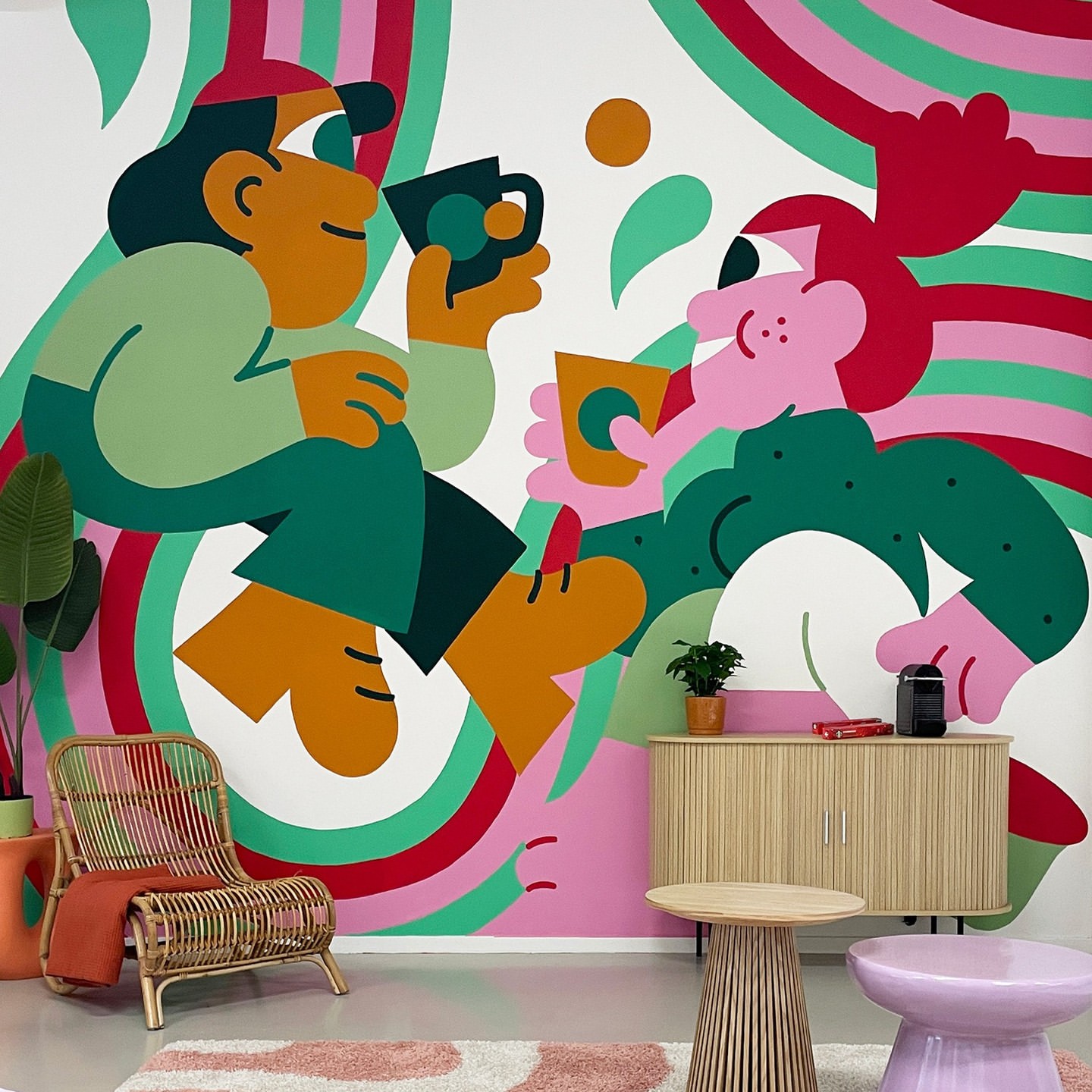 Colorful graphic art of two people enjoying coffee indoors, beside wicker chair and end tables against wall with painted stripes.