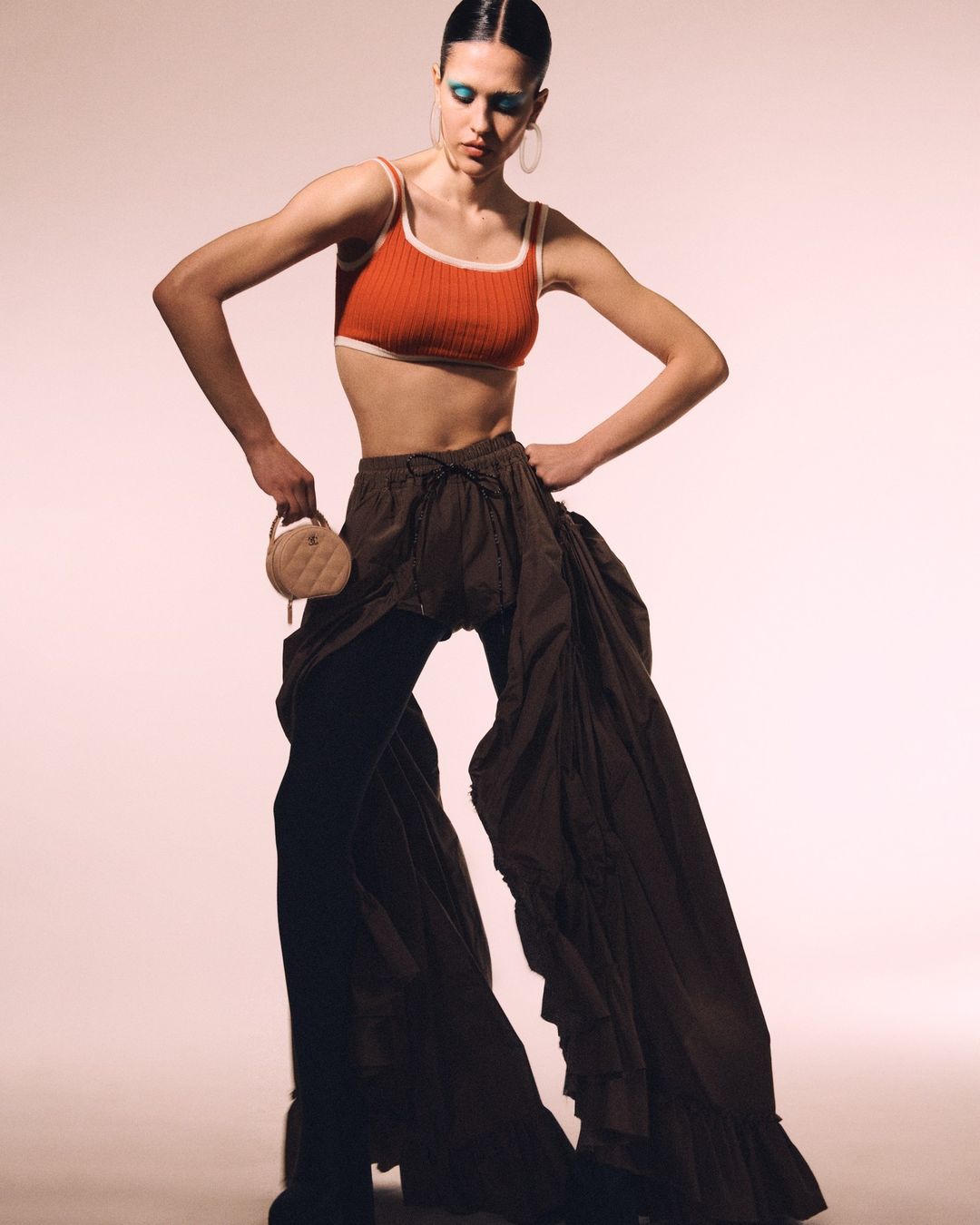 Fashionable woman posing in an orange crop top and dark flowing trousers, complemented by blue eyeshadow and a round purse.