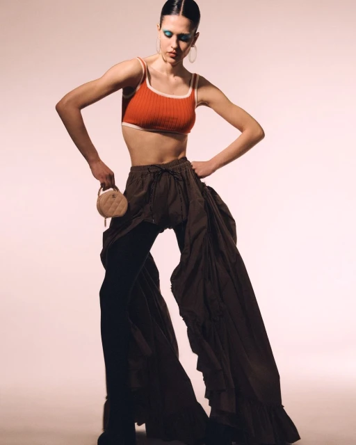 Fashionable woman posing in an orange crop top and dark flowing trousers, complemented by blue eyeshadow and a round purse.