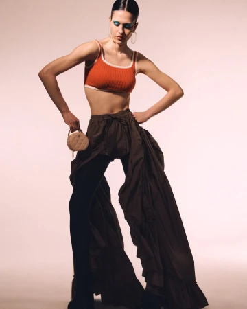 Fashionable woman posing in an orange crop top and dark flowing trousers, complemented by blue eyeshadow and a round purse.