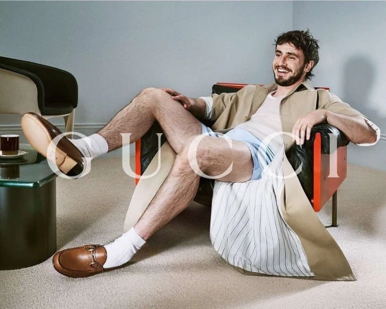 Man seated comfortably in a leather armchair wearing a beige coat, white t-shirt, light blue shorts, white socks, and brown loafers.