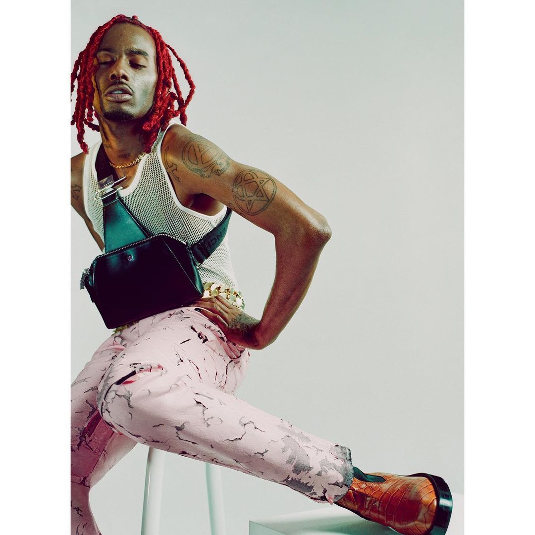 Man with red dreadlocks poses on a stool, wearing a white tank top, leather shoulder bag, patterned pants, and brown dress shoes.
