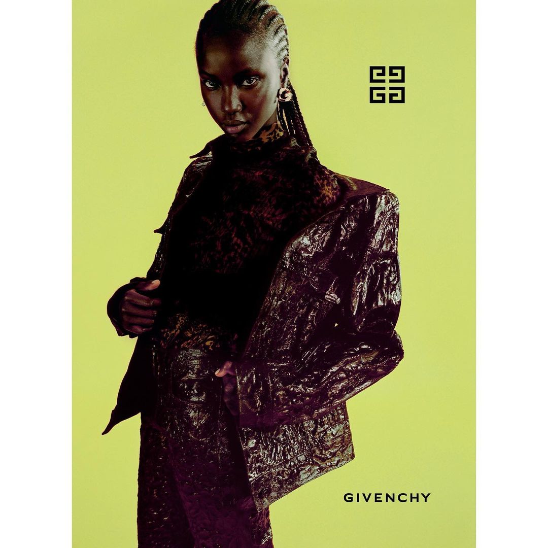 Fashion model poses in a dark glossy jacket and dress against a light green backdrop for a Givenchy advertisement, captured in studio.
