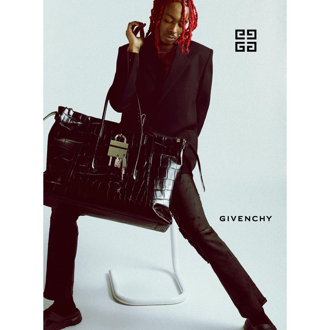 An adult male model sits on a stool next to a black leather handbag in a studio advertisement, featuring the Givenchy brand logo.